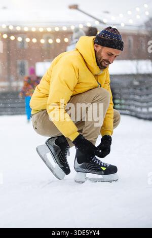 Caucasian young man tying black skates on ice rink in snowy winter day  Stock Photo