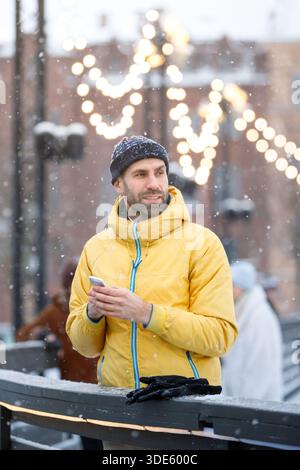Middle aged man standing near fence on ice rink and using smartphone, outdoors at snowy day Stock Photo