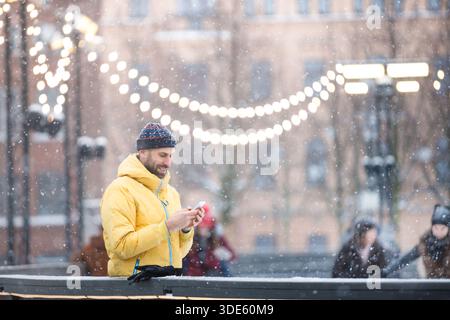 Middle aged man standing near fence on ice rink and using smartphone, outdoors at snowy day Stock Photo