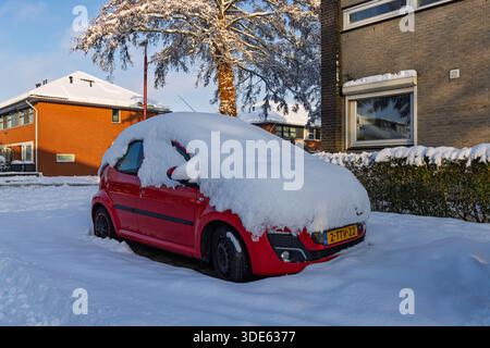 A red car parked on a residential street in the Netherlands completely covered in a thick layer of fresh snow on a sunny winter day.  Utrecht Netherla Stock Photo