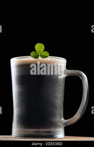 A glass of dark beer with a green shamrock on top on black background. The beer is cold and the glass is frosted Stock Photo