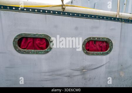 Red curtains draped behind oval windows on the side of a fishing boat Stock Photo