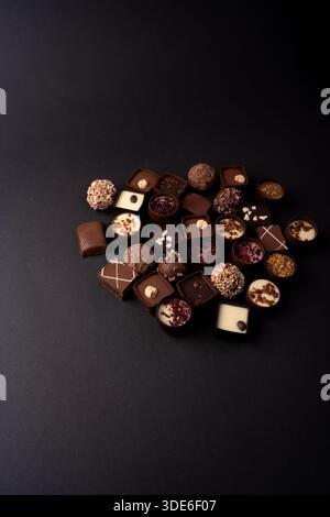 assorted artisanal chocolate pralines and truffles clustered on a matte black tabletop, moody studio lighting highlighting glossy ganache, nut topping Stock Photo
