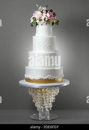 Elegant four-tier white wedding cake decorated with lace-style piping, floral detailing, gold accent band, and a monogram, topped with pastel sugar fl Stock Photo