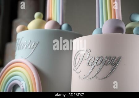 A creative joint birthday cake display with two separate tiers, silver script names, and a tall pastel rainbow bridge, presented on a white pedestal s Stock Photo