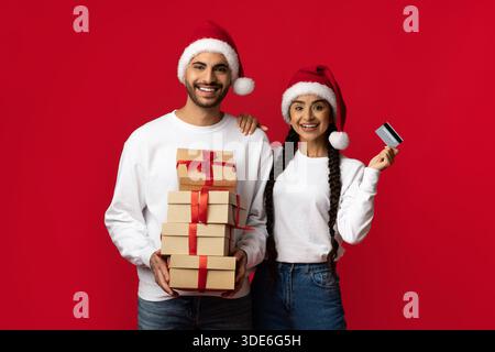 Joyful Holiday Couple Celebrating Christmas With Gifts and a Credit Card in Front of Red Backdrop Stock Photo