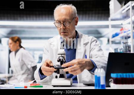Senior lab manager checks product surface for tiny defects using microscope. Elderly research facility professional ensures manufacturing standards, examines small parts with optical device Stock Photo