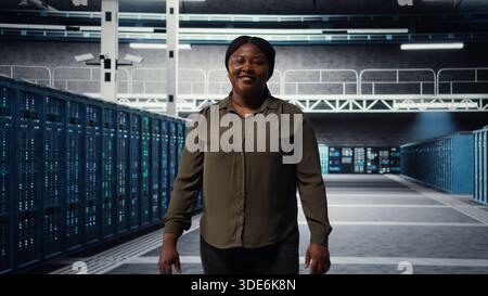 Portrait of smiling admin walking in data center, overseeing large scale IT infrastructure to maintain seamless operations. Happy server farm employee pleased by increased uptime, camera A Stock Photo