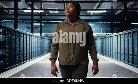 Supervisor in data center overseeing large scale IT equipment to maintain seamless operations. IT expert walking between server room rows, doing network management for increased uptime, camera A Stock Photo