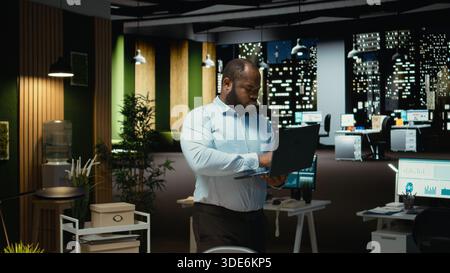 Black guy examining archived documents before transcribing on pc notebook, notetaking and extracting the main points from reports on laptop. Analyst documenting after hours late at night. Stock Photo