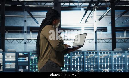 Software developer in data center using laptop to coordinate deployment of new server architecture. Server hub IT professional using notebook to do rollout of cybersecurity framework, camera B Stock Photo