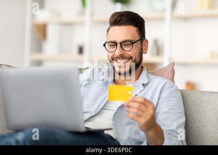 Easy payment and cashback concept. Smiling young man holding credit card and using laptop computer, sitting on couch Stock Photo