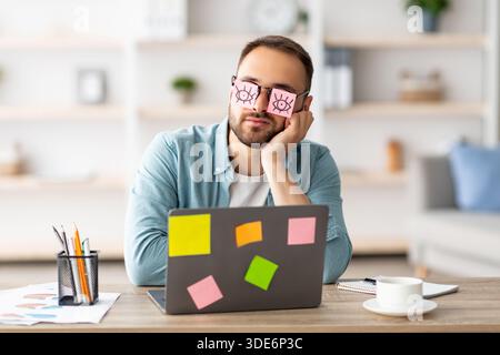 Tired young man having stickers with open eyes drawing on glasses, sleeping near laptop, pretending to work at office Stock Photo
