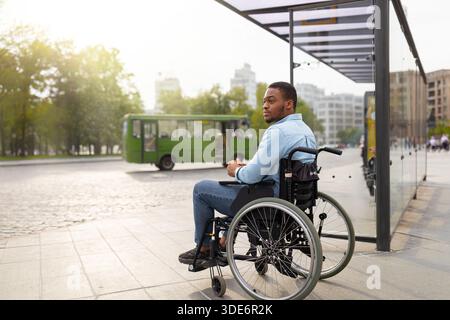 Young disabled black man in wheelchair waiting for public transport on bus stop, having difficulty traveling around city Stock Photo