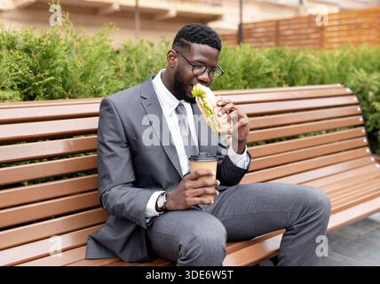 Young african american office worker having lunch outdoors, drinking coffee and eating sandwich, sitting on bench Stock Photo
