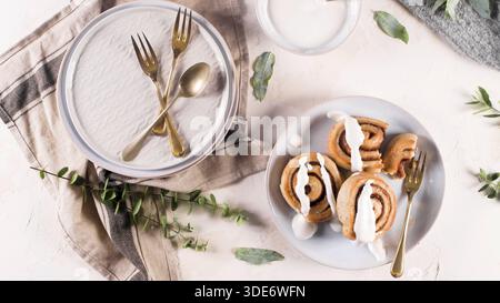 Freshly baked cinnamon rolls drizzled with white icing rest on ceramic plates. The scene features elegant gold cutlery, linen napkins, and green eucal Stock Photo