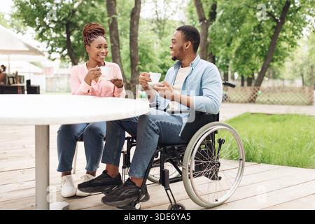 Millennial black guy in wheelchair having date with beautiful woman at outdoor cafe, enjoying coffee and conversation Stock Photo