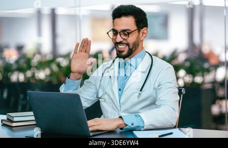 Doctor Engages in Virtual Consultation While Smiling and Waving at the Camera Stock Photo