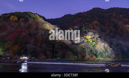 A nighttime view of the Katsura River in Arashiyama, Kyoto showcases illuminated autumn foliage along the riverbanks. The photograph captures a serene Stock Photo