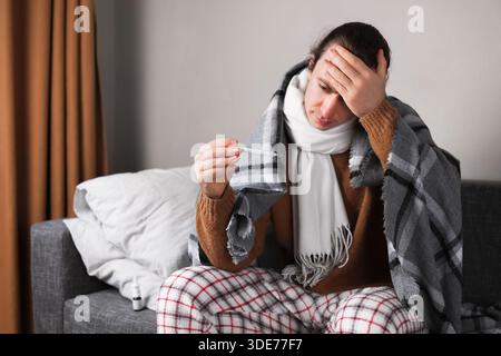 Sick man in blanket checking temperature with thermometer and touching forehead on sofa. High quality photo Stock Photo