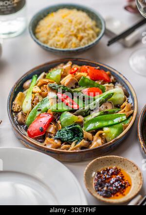 Stir-fried rice cakes with mixed vegetables served on a well-set dining table, accompanied by a bowl of rice and chili oil. Stock Photo