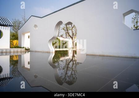 Perspective view of a white architectural wall with cloud and hexagonal shaped openings reflecting in a water feature. The image conveys geometric des Stock Photo