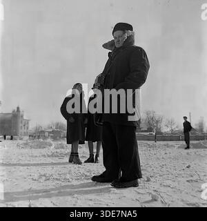 Slaviansk, Donetsk region, Ukraine, USSR. Early 1950s archival winter photo. A portrait of a man standing on the frozen surface of Lake Liman near the Red Metalworker plant. He is dressed in a dark coat with a fur collar and a warm fur hat. A leather camera case hangs around his neck, indicating he is a photographer or hobbyist. In the background, two women and a skater are visible near the city buildings. This square medium format image captures the atmosphere of leisure and personal hobbies in a peaceful Donbas before the war. Stock Photo