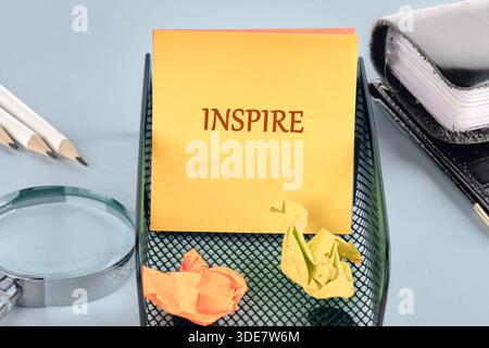 A yellow sticky note with the word INSPIRE is displayed in a holder. Pencils, a magnifying glass, and crumpled papers create an evocative scene. Stock Photo