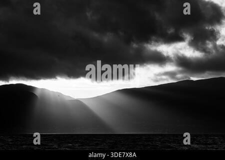 Black and white sunrise light breaking through heavy cloud over the Carneddau mountains, seen across the Menai Straits Stock Photo