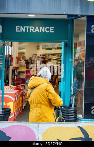 older woman standing at the entrance to a high street store wearing a yellow winter coat. Stock Photo