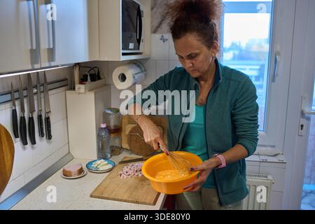 Woman whisks eggs in orange bowl while preparing omelette for breakfast in home kitchen Stock Photo