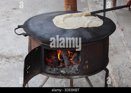 A thin sheet of dough for traditional Turkish Gözleme pastry being cooked on a circular metal sac oven over an open wood fire Stock Photo