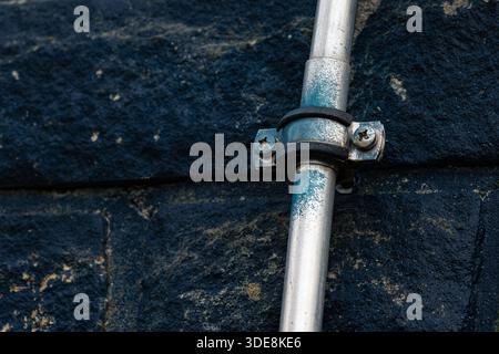 A metal pipe is attached to a dark stone wall. The pipe has a shiny finish and is held in place with metal clamps and screws. This scene is common in Stock Photo