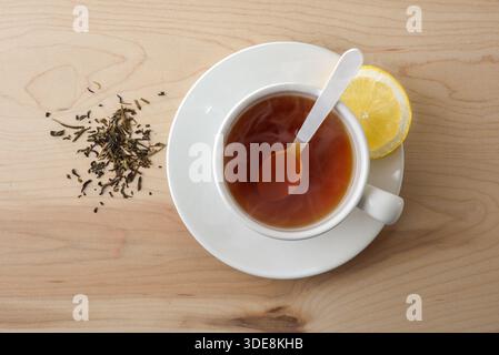 Background with detail of black tea with lemon in a white ceramic cup on a wooden table and dried black tea leaves. Top view. Stock Photo
