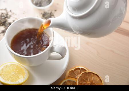 Details of serving black tea from a ceramic teapot into a white ceramic cup with a slice of lemon on a wooden table with ingredients and tea leaves. E Stock Photo
