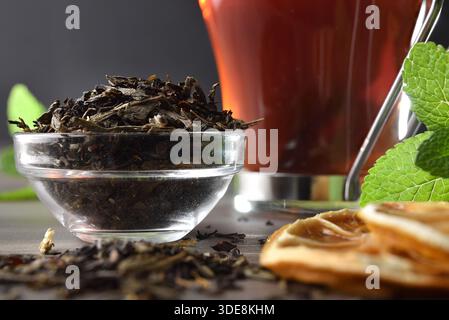 Detail of bowl full of dried tea leaves and black tea infusion in a glass cup  with orange slices and mint on a wooden table and dark isolated backgro Stock Photo