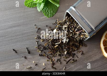 Detail of dried black tea leaves spilling out of a metal container on a dark wooden table with mint leaves and a slice of dehydrated lemon. Top view. Stock Photo