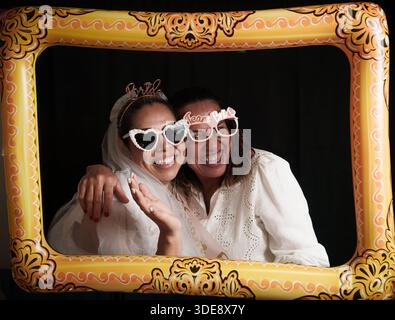 Two women wearing sunglasses and holding hands. One of them is wearing a wedding dress. The frame is yellow and has the word bride on it Stock Photo