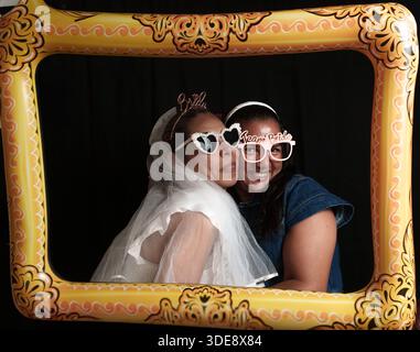 Two women wearing sunglasses and glasses are posing for a picture in front of a yellow frame. The frame has the words "Beauteous" written on it Stock Photo