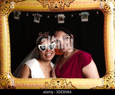 A woman wearing a white dress and sunglasses is posing for a picture. A woman in a red dress is standing next to her Stock Photo