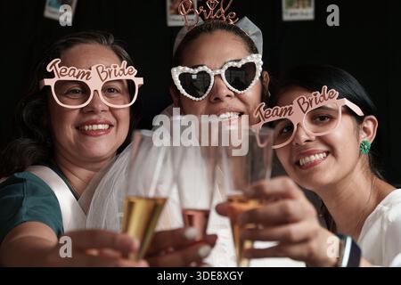Three women are wearing team bride glasses and holding glasses of champagne. They are smiling and toasting to the occasion Stock Photo