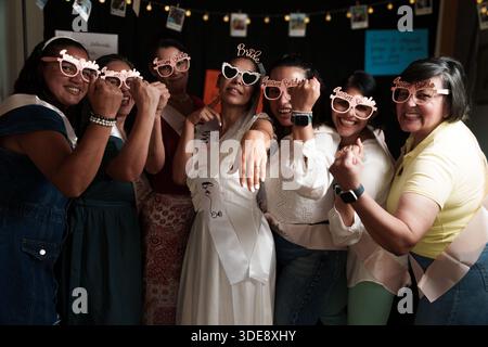 A group of women are posing for a picture with sunglasses on. One of the women is wearing a white dress and a white tiara Stock Photo
