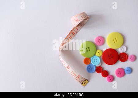Top view of various colorful buttons next to a curled tailor measuring tape on a white surface. Creative sewing and craft concept. Stock Photo