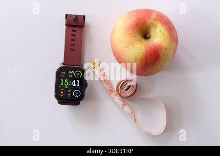 Top view of a digital smartwatch, a fresh red apple, and a tailor measuring tape on white background. Concept of healthy lifestyle and dieting. Stock Photo