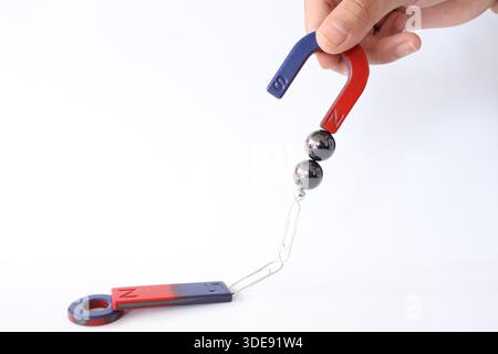 A hand holding a horseshoe magnet attracting steel balls and paperclips connected to a bar magnet. Science education concept on white background. Stock Photo