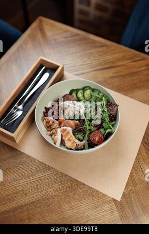 A bowl filled with fresh salad, grilled chicken, cucumbers, cherry tomatoes, and herbs sits on a wooden table. A wooden cutlery holder is nearby with Stock Photo