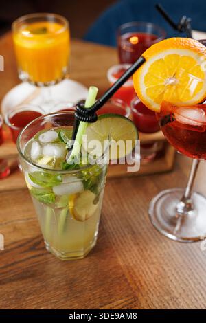 A group of colorful drinks is displayed on a wooden table at a bar. A mojito with fresh mint and lime stands in front, while other cocktails and juice Stock Photo