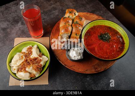 A table displays a bowl of Ukrainian borscht soup, dumplings topped with fried onions, soft bread rolls, and a glass of berry drink. Various flavors c Stock Photo