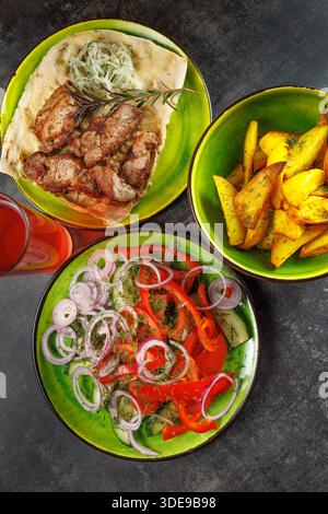 A table displays a selection of grilled meat with flatbread, fresh salad with onions, and crispy potatoes in colorful bowls. A refreshing drink is als Stock Photo