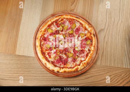 A round pizza is placed on a wooden surface. The pizza is topped with slices of ham, green peppers, and olives. It is ready to be enjoyed. Stock Photo